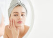 THE SKIN CENTER: Collagen blog. IMAGE: A white woman wearing a bathroom cap draws her hand to her face, examining her face in the mirror.