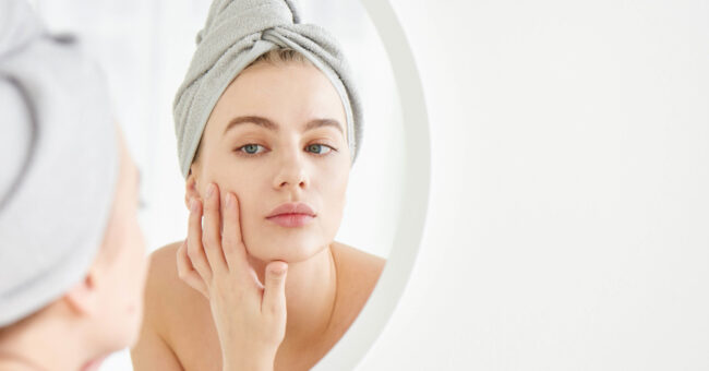 SOUTHGATE_SKIN_COLLAGEN THE SKIN CENTER: Collagen blog. IMAGE: A white woman wearing a bathroom cap draws her hand to her face, examining her face in the mirror.