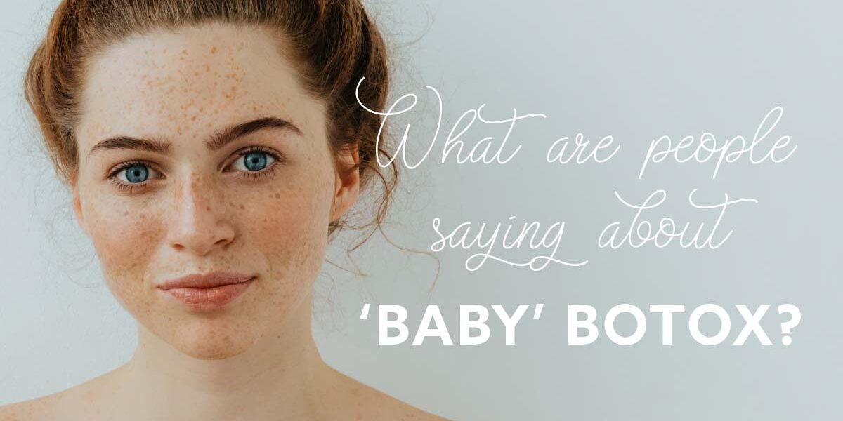The Skin Center_Baby Botox A red-headed young woman with blue eyes, freckles, and pink lips, stands in front of a baby blue background. Text reads in white cursive:"What are people saying about," followed by white block letters: "'BABY' BOTOX?"